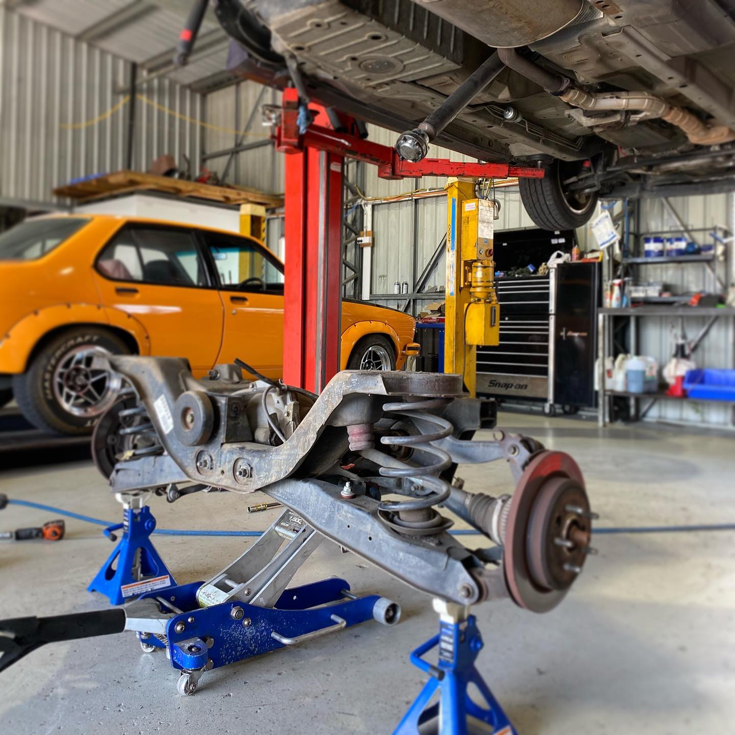 Rear suspension and subframe assembly out of a car on jack stands, an orange Torana on the hoist behind