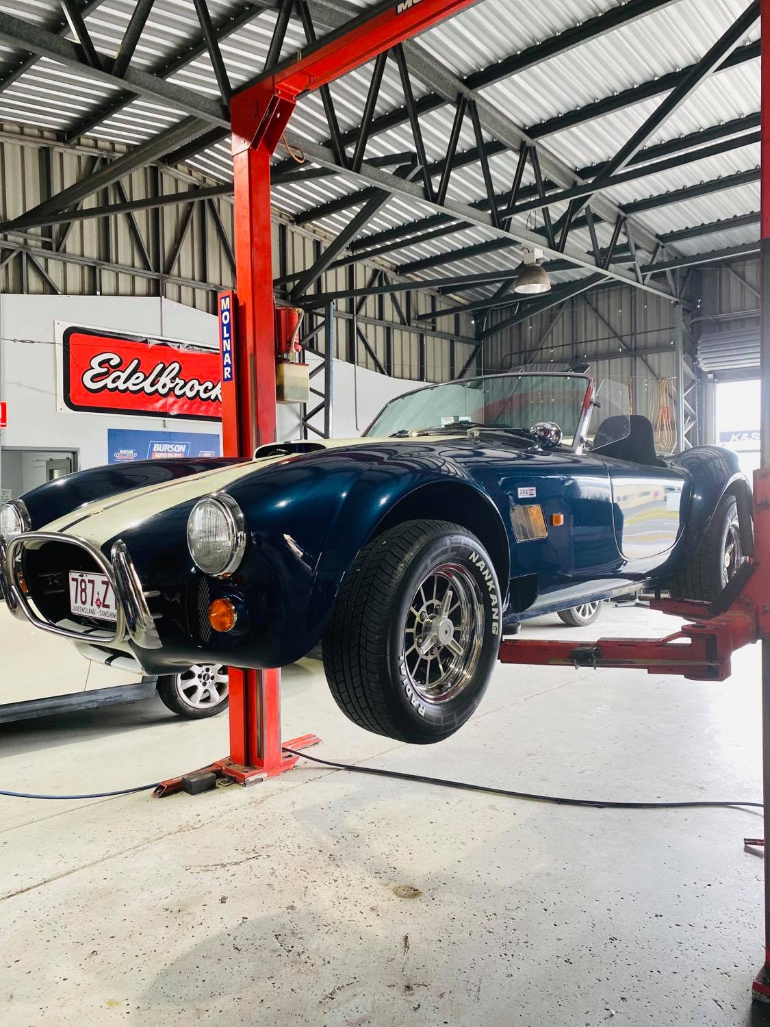 A dark blue AC Cobra replica raised on the two post hoist inside the Bowser workshop